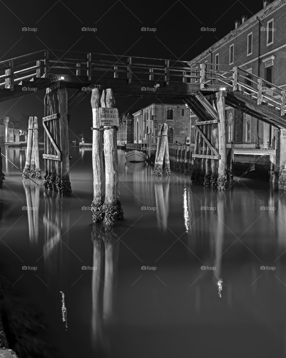 Night Enchantment. Nocturne of the access canal to the Arsenale of Venice, with ancient doors, poles immersed in the water and wooden bridge that are reflected in a calm water like a mirror.
