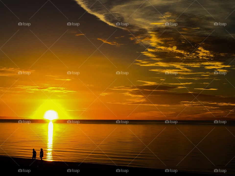 Sun rising over the horizon and reflection on calm ocean, couple walking toward the sunrise on the beach, brilliant hues of oranges and yellows