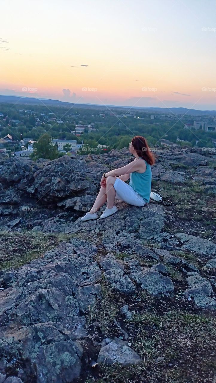 girl looks into the distance, meditation, contemplation nature, view from the mountain to the city,  summer, evening, calmness, peace, light breeze, the girl is sitting on the grass