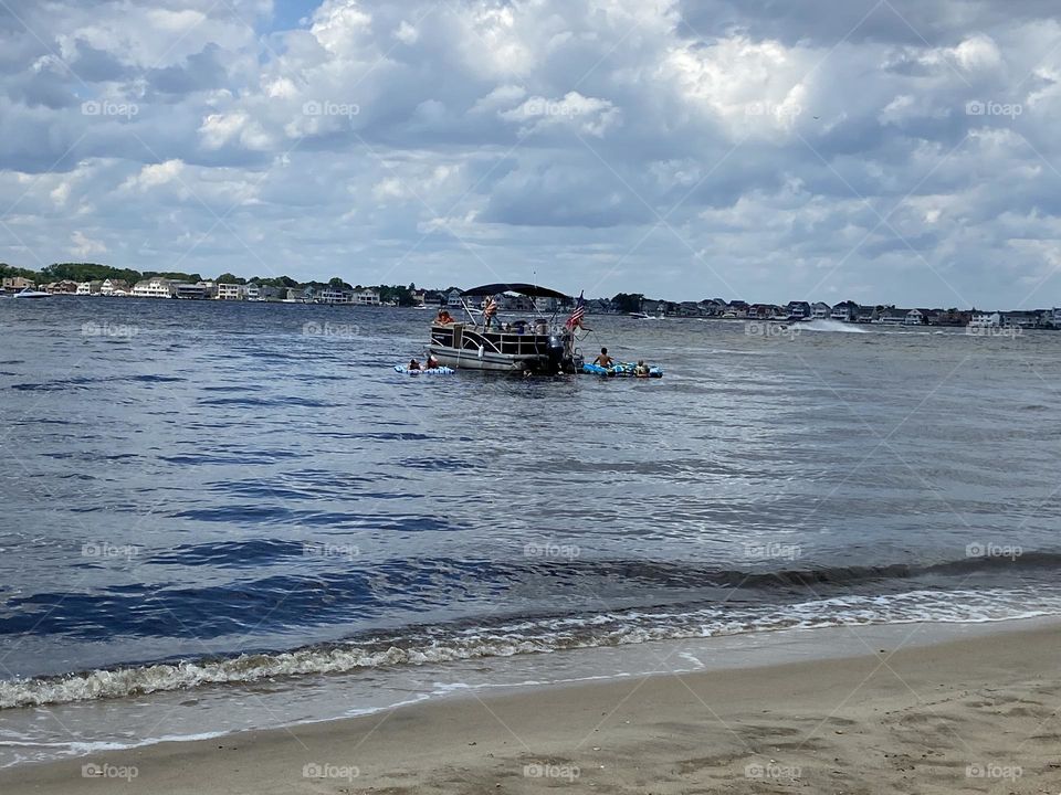 People boating on Silver Bay viewed from the beach at Cattus Island Park in Toms River, NJ. Waves lap at the edge of the sand and clouds dot the blue sky. The water is beautiful whether you are on it or just watching from the sand. 