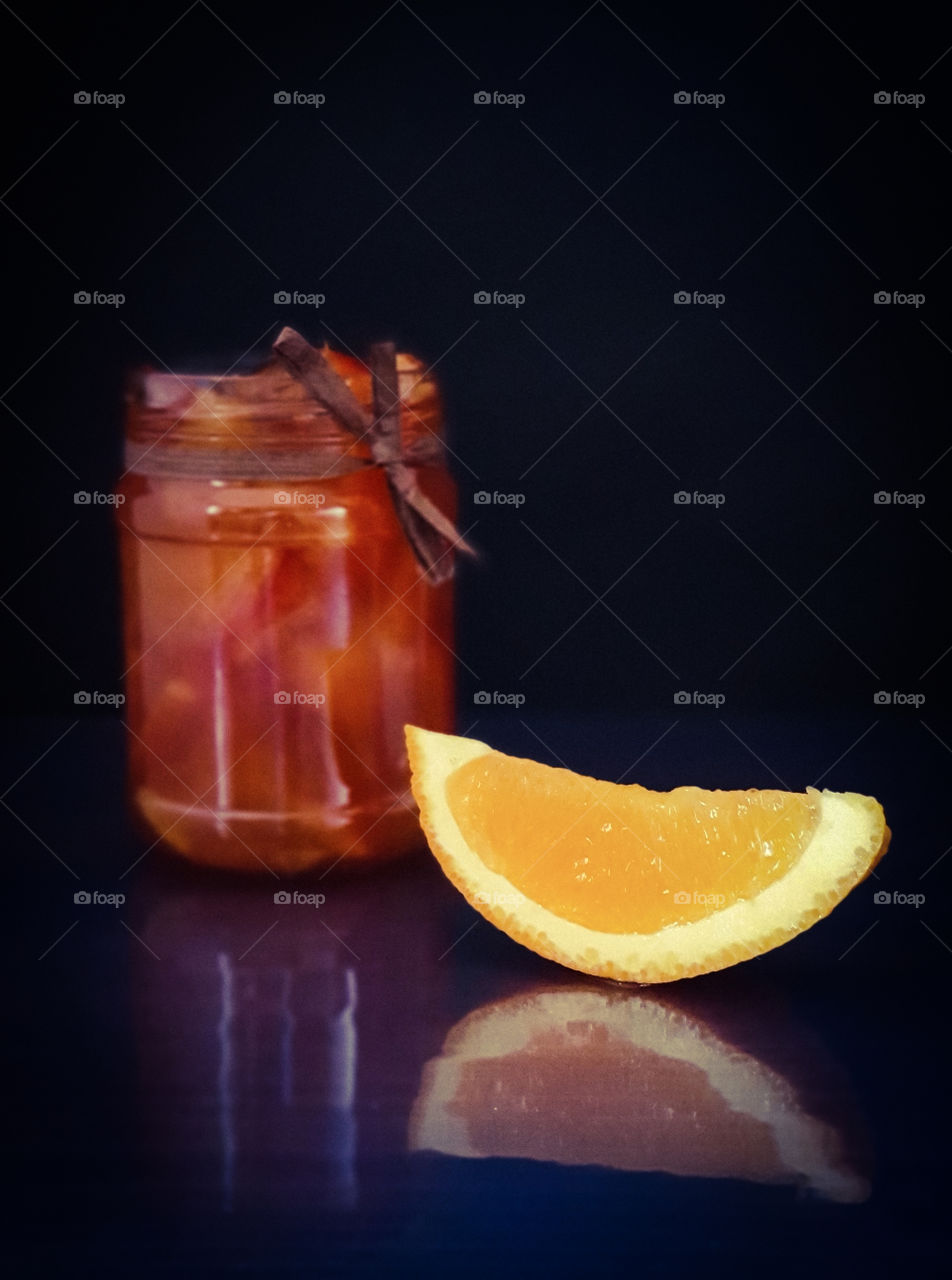 Autumn still life with reflection. Jar of orange jam tied with a decorative ribbon with a slice of juicy orange on a dark background with reflection