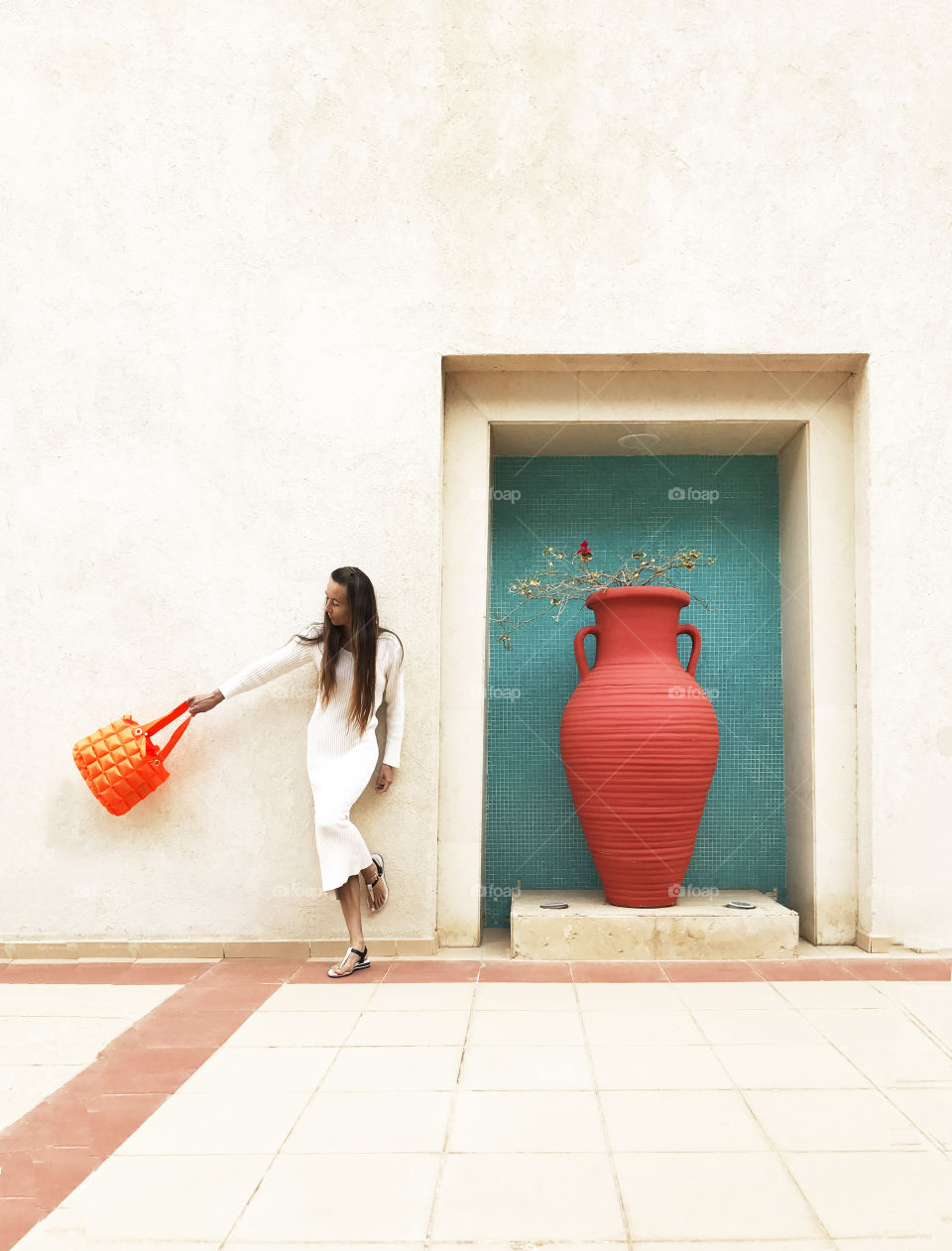 Young woman wearing white dress and holding a bright orange purse standing nearby huge orange jar