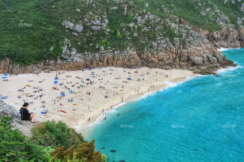 Looking From above . A lone girl sitting on cliff tops admiring the view of a busy, sandy beach.