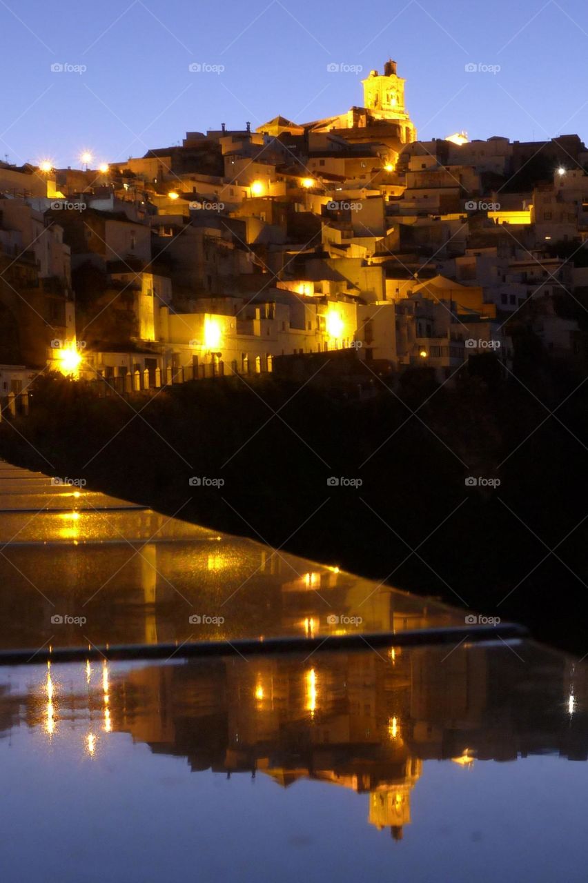 In Andalusia, a small village lights up at nightfall. In the foreground, the reflection of the lights.