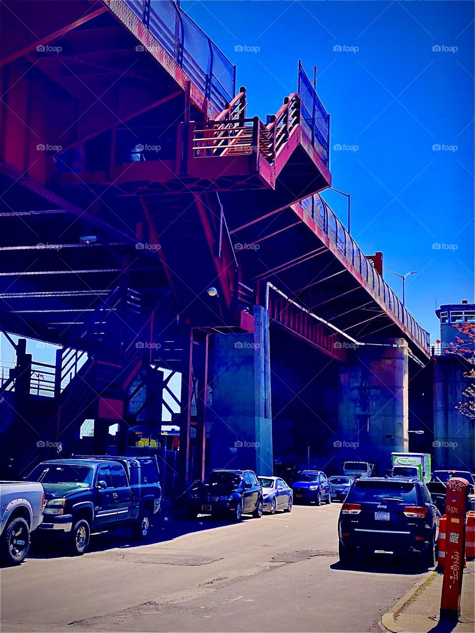 The parking lot beneath the „Pulaski Bridge“ contains a red metal staircase leading up to the pedestrian walkway across to „Greenpoint“, Bklyn. The industrial design of the bridge & the stairs is functional & attractive. 2022. Hypnotic Productions
