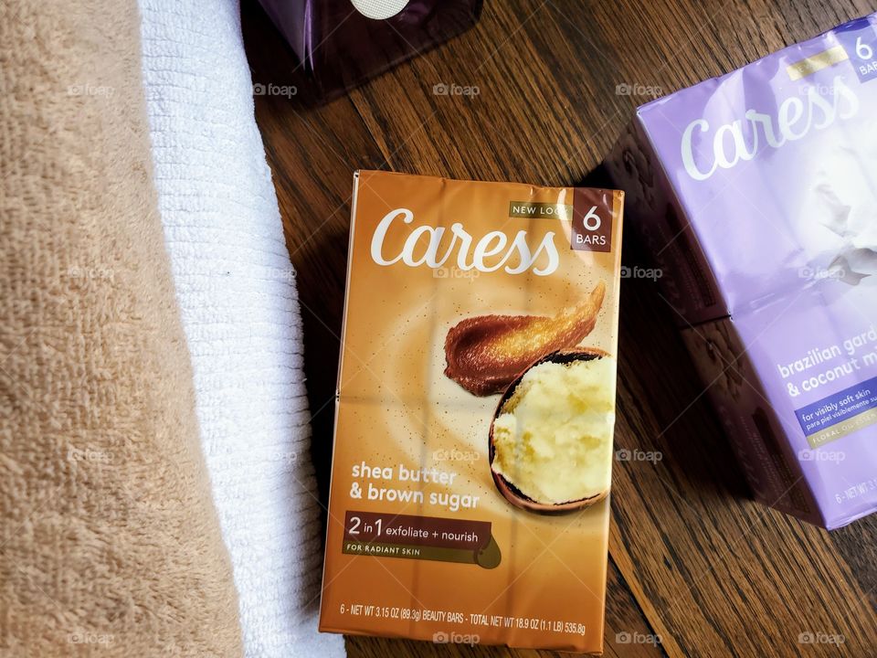 The new look Caress bar six pack of shea butter & brown sugar in the center and brazillian gardenia & coconut milk on the right. Towels on the left. All on a wood surface.