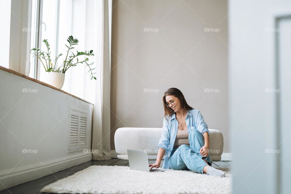 Young woman in jeans working on laptop sitting on carpet at home, view from doors