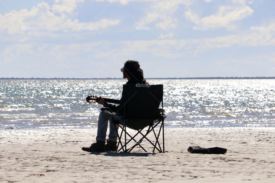 Soulful musician playing guitar on beach surrounded by glistening ocean late day golden hour