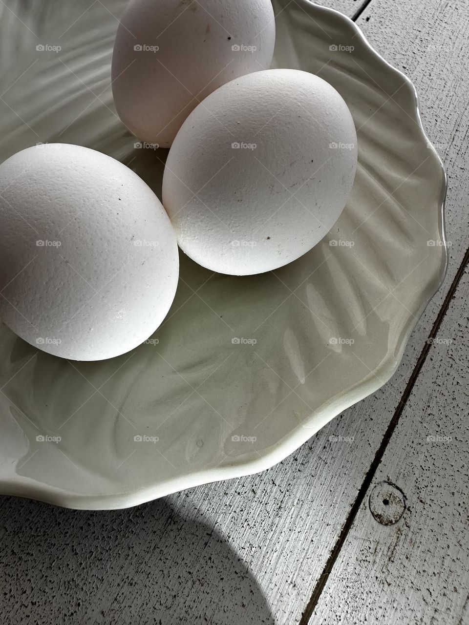 iPhone 14 Pro photo of three white fresh chicken eggs on a ceramic plate with leaf pattern on a whitewashed wooden board surface