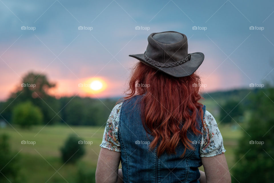 Woman with red hair and hat in head looks at sunset in the distance. Rear view.