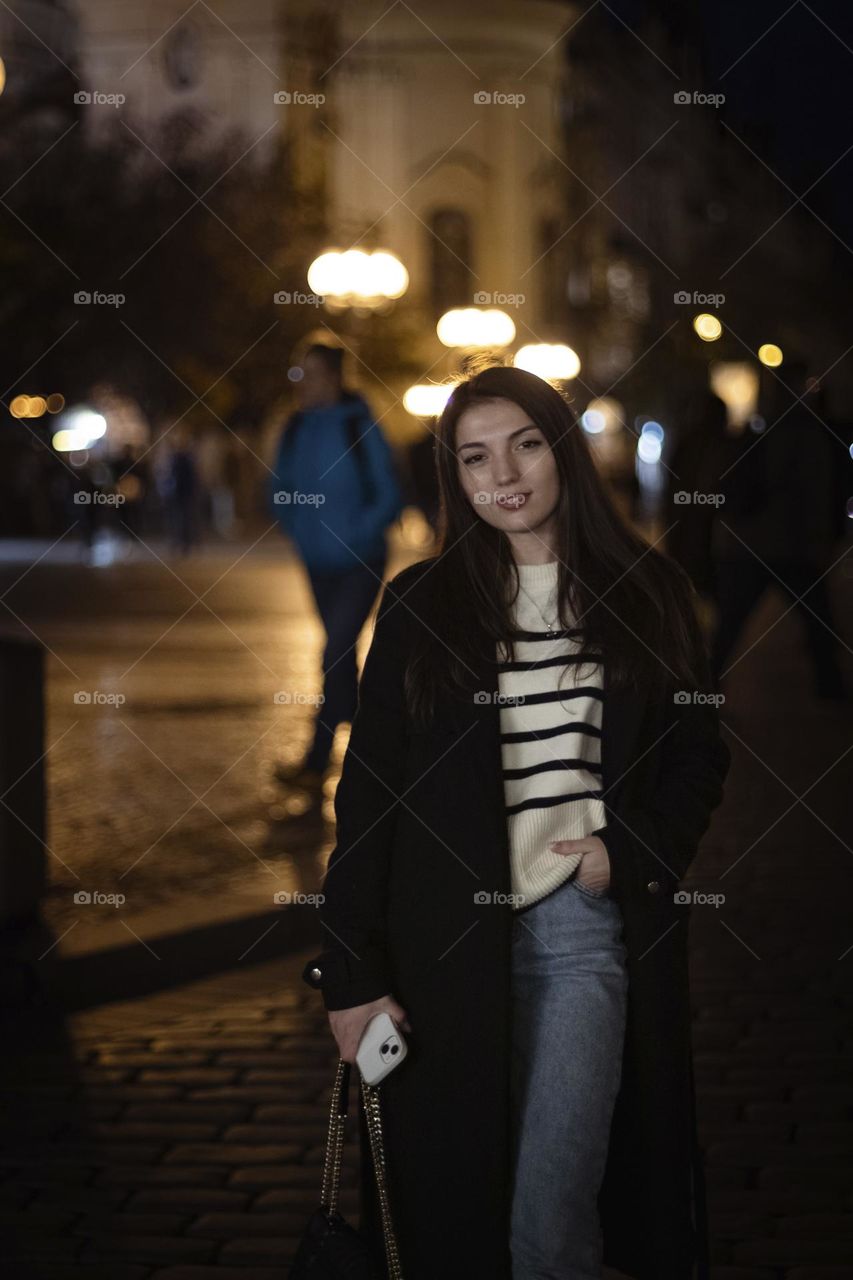 Portrait of a beautiful young caucasian brunette girl with long flowing hair, black coat with a sweet smile on her face looking at the camera and walking around the ancient city of Prague in the Czech Republic, close-up side view.