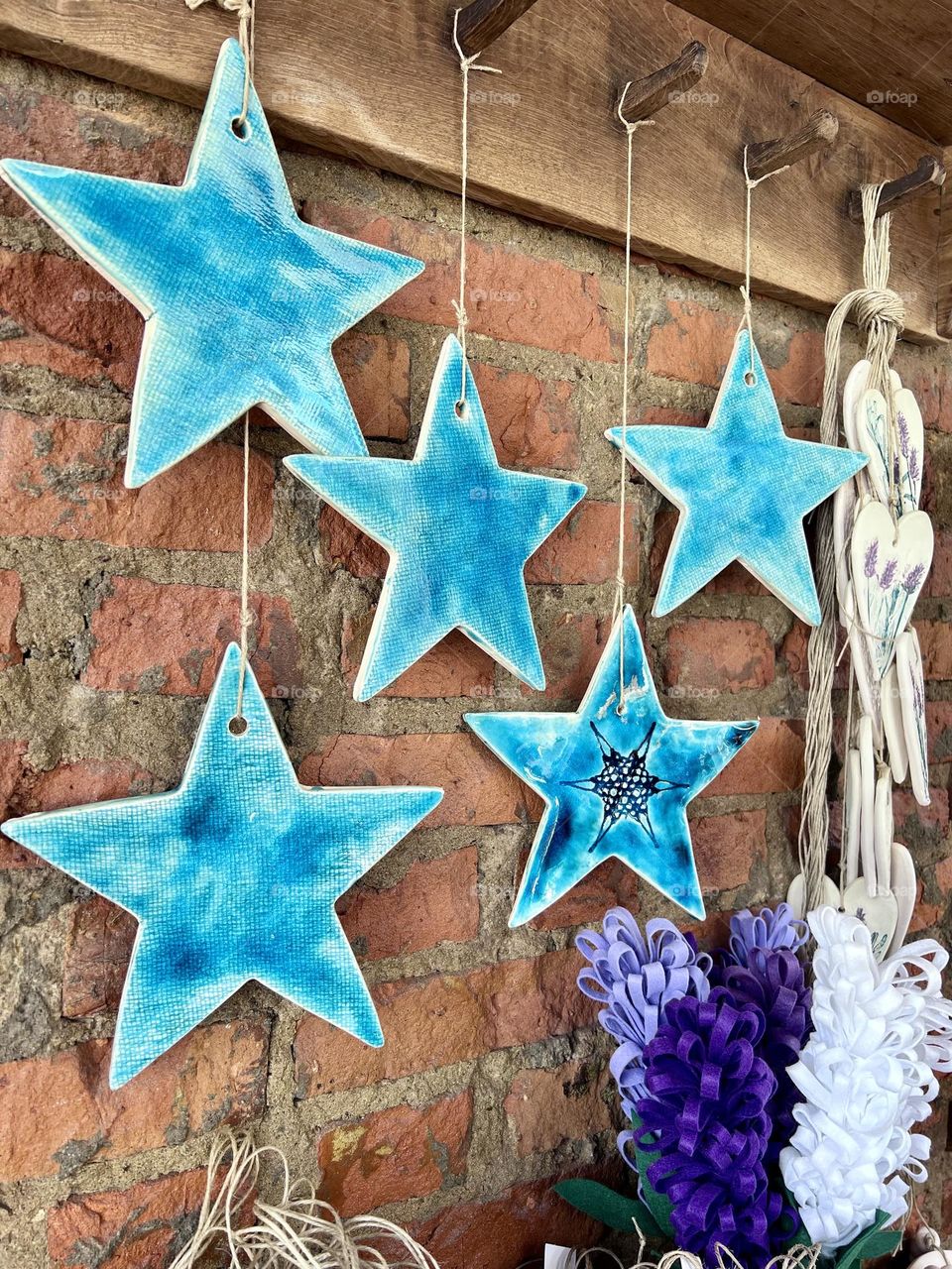 Uma encantadora exibição de estrelas de cerâmica azuis penduradas em uma parede de tijolos rústicos, complementadas por detalhes em vime e decoração floral roxa, criando um ambiente caprichoso e artístico.