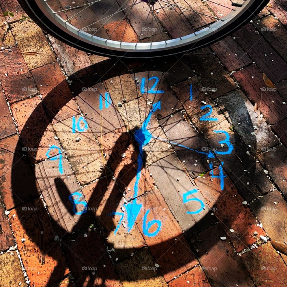 A creative shot of a sidewalk chalk painting of a clock using the shadow of the spokes of a bicycle tire