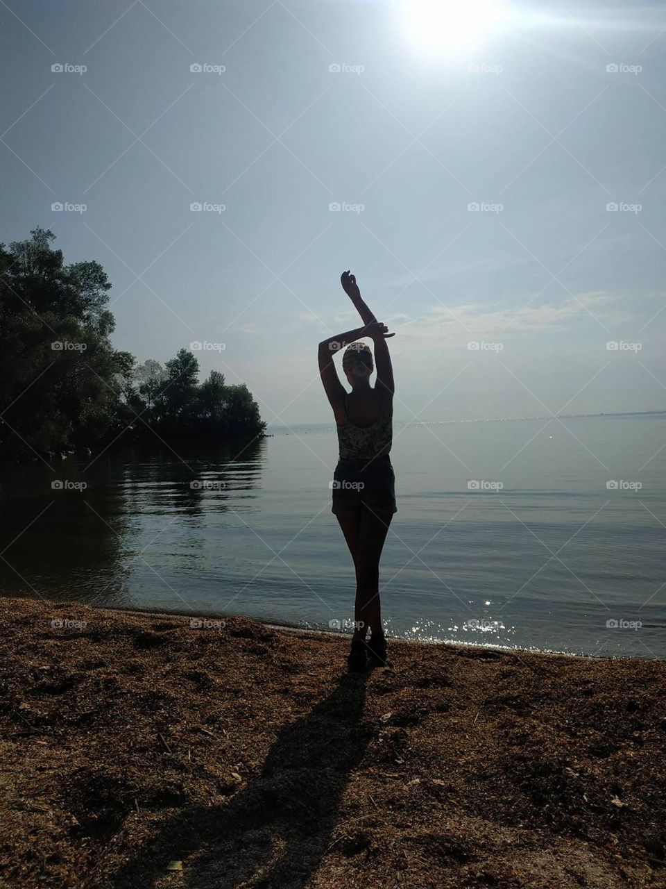 Woman on the beach, near the water. Figure shadow, sunny day, woman's silhouette. Trees shadows on the water. Girl with hands up, meditation pose