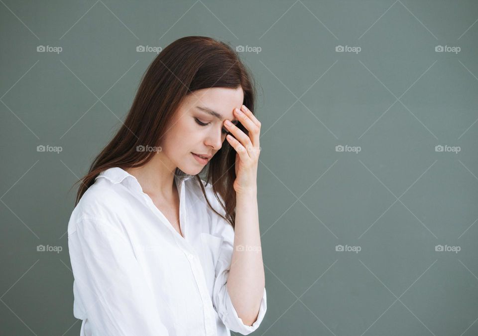 Young unhappy woman with long hair in white shirt on grey background, negative emotion