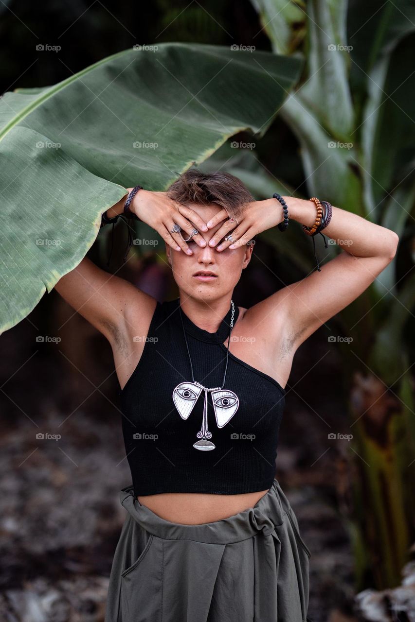 Portraut of a beautiful woman with short haircut starting among banana trees. Eyes closed with her hands. Stylish accessories