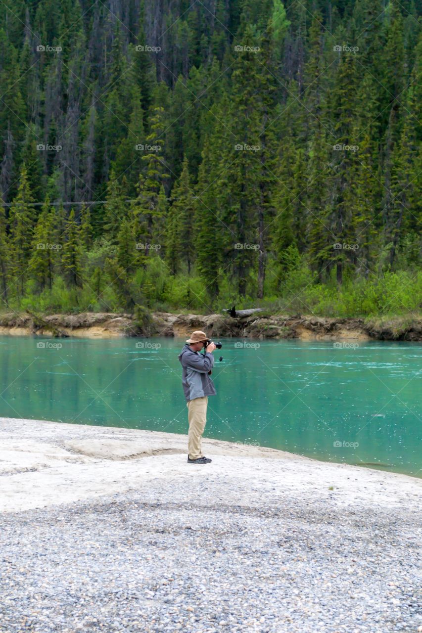 Male photographer traveller tourist beside green water river in Canada's Rocky Mountains shooting a photograph 