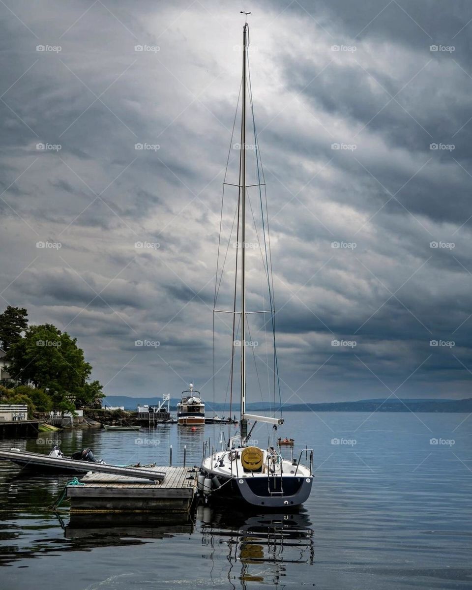 The composition of the sailboat with its striking yellow sail provides a beautiful contrast against the surrounding environment. The overall atmosphere suggests a quiet day on the lake, perfect for relaxation and contemplation.