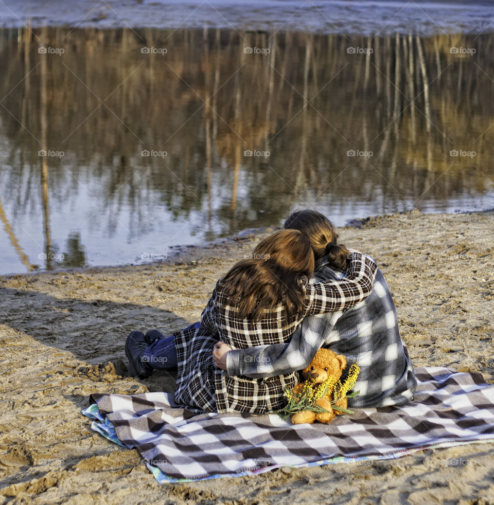 Young couple sit and hug on the blanket on the sand next to a lake