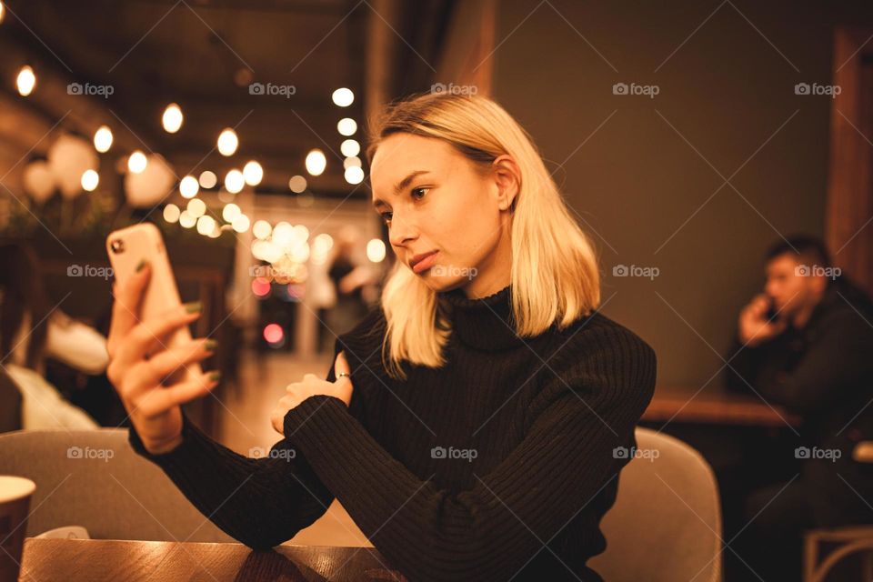 A young woman sits in a cafe and speaks on the phone. Happy blonde is resting in a cafe. Portrait photo of a woman in a black sweater