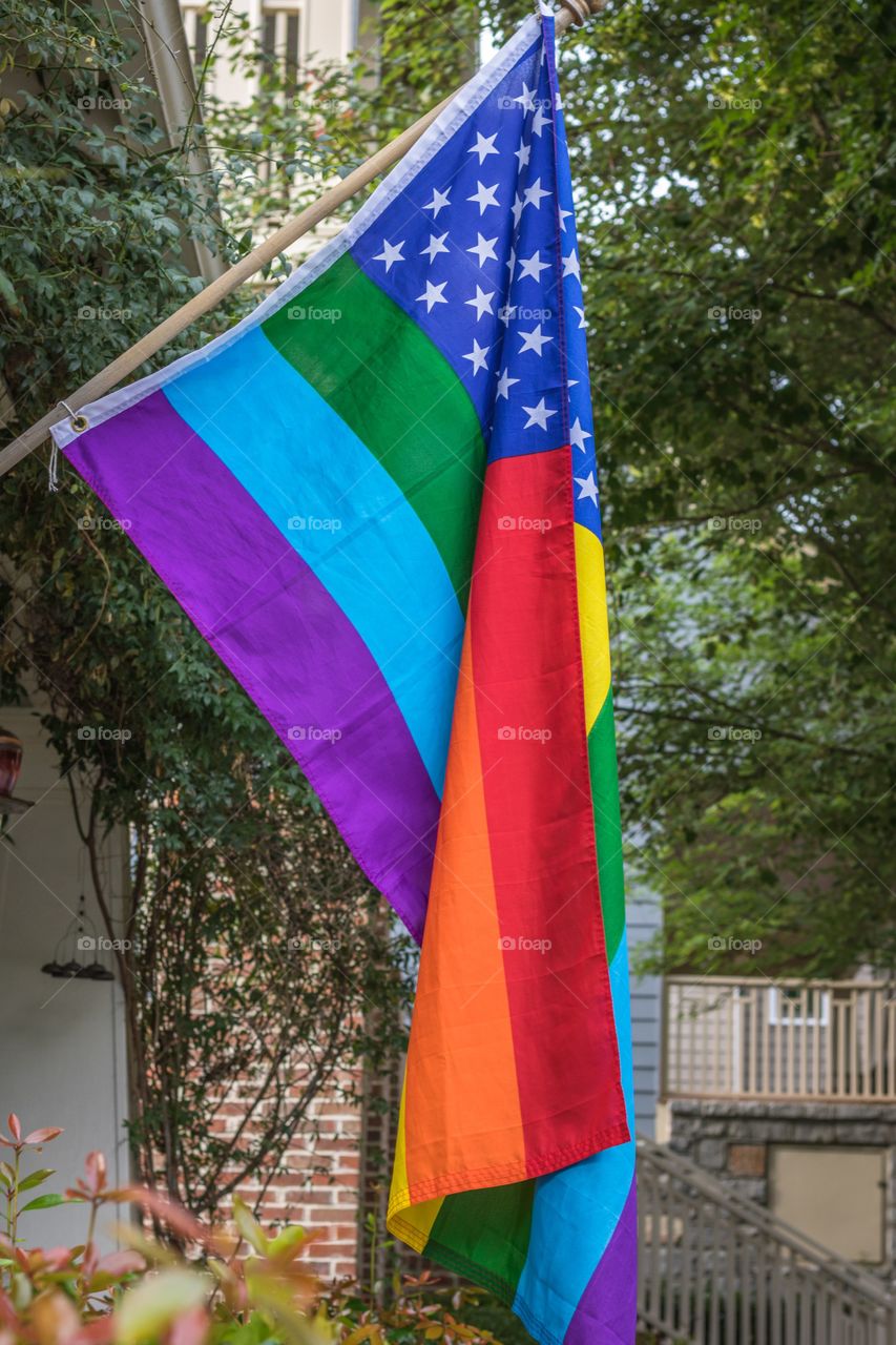 Vertical photo of a mixed American and LGBTQ Pride flag hanging in a residential neighborhood with green trees in the background