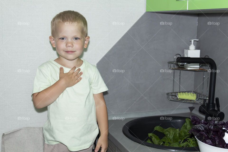 A small child washes fresh, green, vegetarian vegetables under the tap in a black sink to prepare salads and other dishes in a homemade gray and green kitchen.
