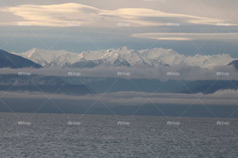 Peaceful ocean with haze over it and snow caped mountains rising above and cloudy sky tops it all 
