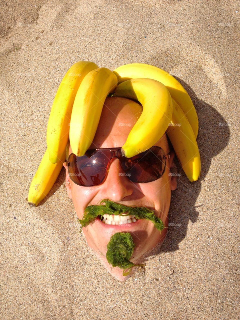 Cheerful man with seaweed and banana buried in sand