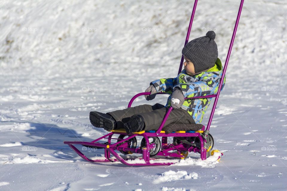 A child with a serious expression on his face in winter clothes jackets, pants, hat and boots in winter on white snow on the street and in the park in nature sledding and playing winter fun.