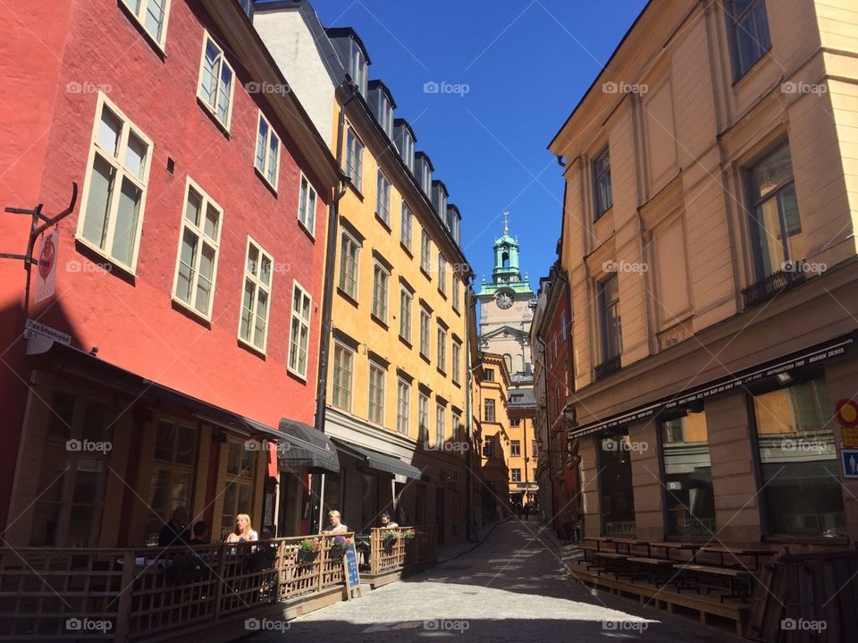 A narrow, Swedish street with patrons eating at a cafe, a church tower is seen in the background of the other colorful buildings. 