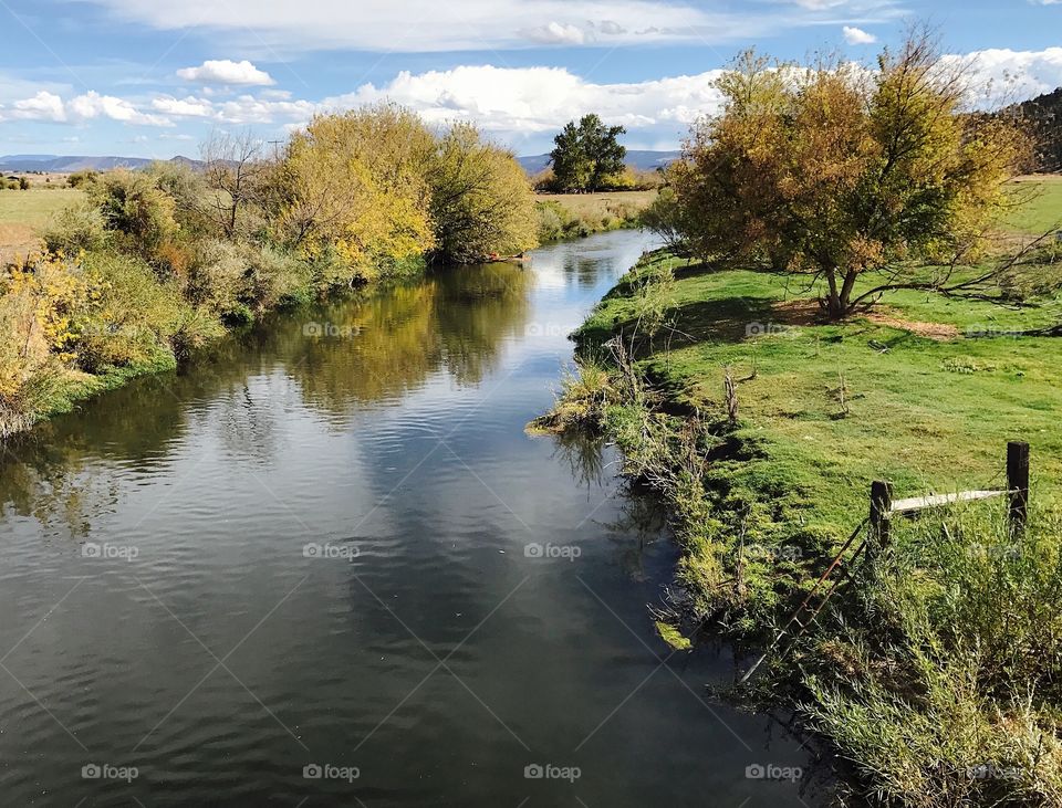 The Crooked River in Central Oregon winds through farmland with colorful trees and clouds reflecting off of its surface on a sunny fall day. 