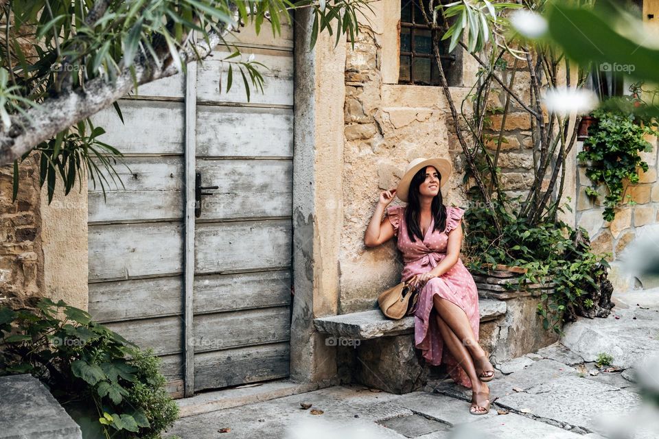 Young woman sitting on stone bench in front of old house in alley full of plants