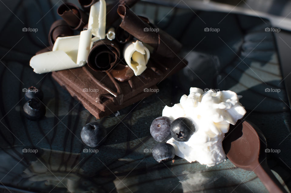 Blueberries and whipped cream on spoon made of chocolate on plate with thin dark chocolate biscuit sandwich draped with sauce and covered with a pile of white and dark chocolate shavings