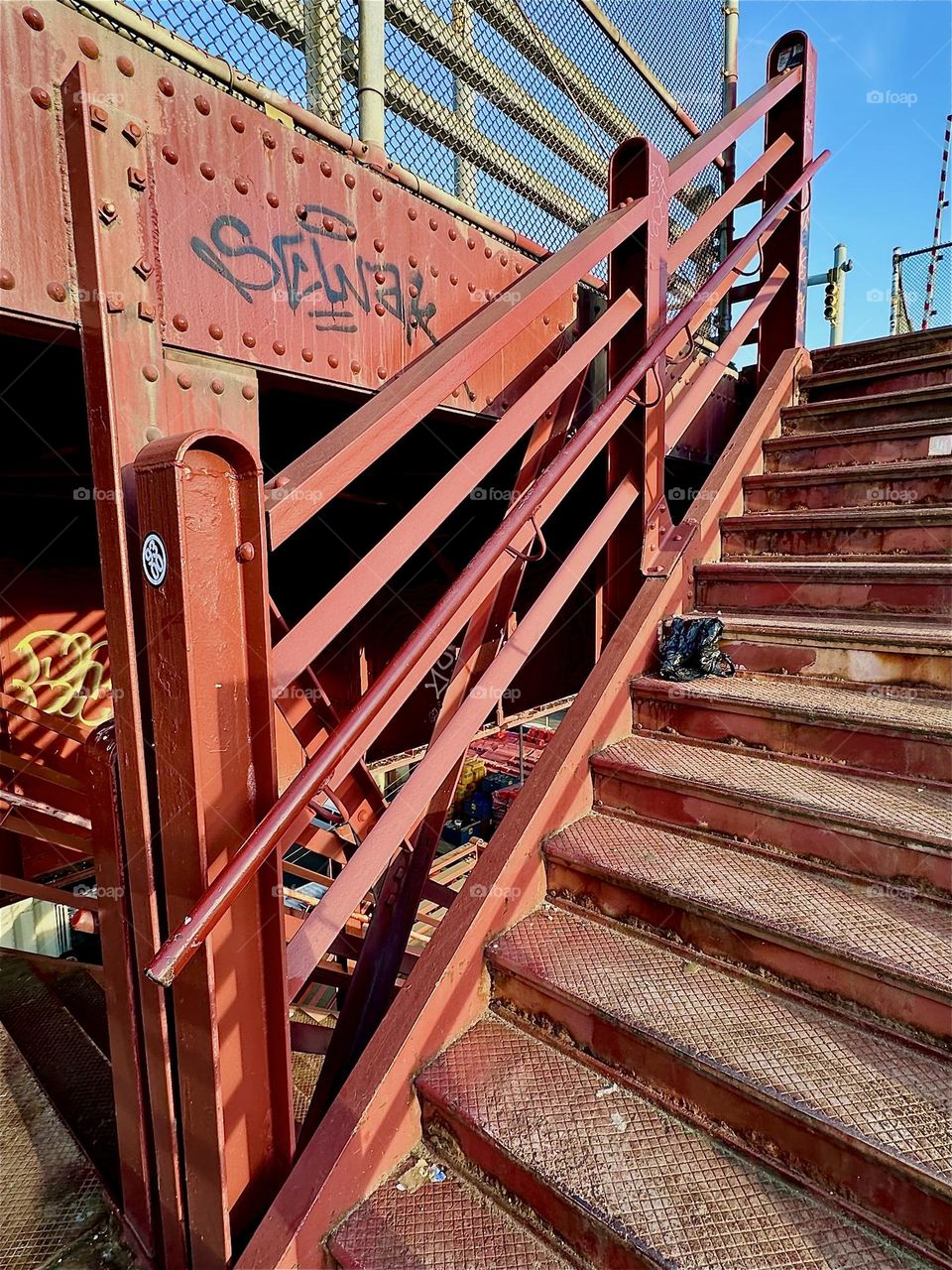 This is the red metal staircase to the pedestrian lane of the “Pulaski Bridge” at “Newtown Creek” in LIC, Queens. The “Bauhaus” inspired strong geometric elements are held together by gravity and heavy duty studs and bolts. 2024. Hypnotic Productions