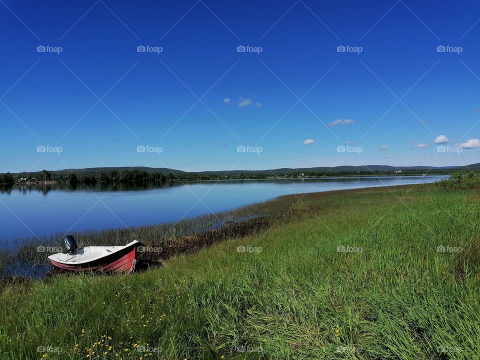 Red old small boat on the bank of the Tornio river. The water is calm, no wind at all. A warm summer day in Ylitornio, Lapland, Finland.