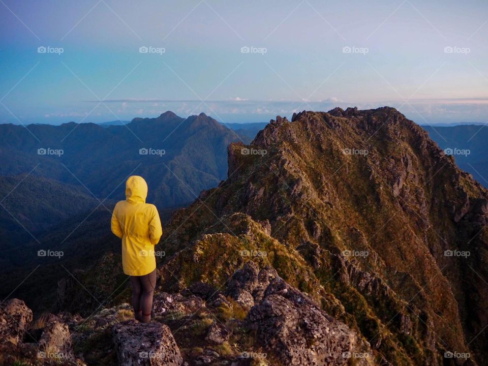 A hiker wearing a yellow jacket stands atop a rocky ridge line watching the sunrise