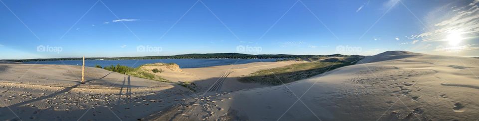 A park organic view of the sand dunes overlooking Silver Lake in Michigan. The sky is a beautiful blue, the tree line a lustrous green, the water a sapphire blue, the sand a sultry tan, and the dune grass a vibrant green. Two shadows of people. 