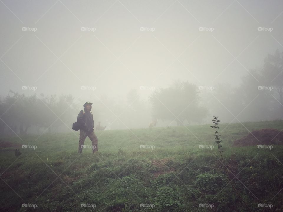 Man in the fog. Fogy day in the field, rain is about to start. Behind him is deer standing and looking at him.