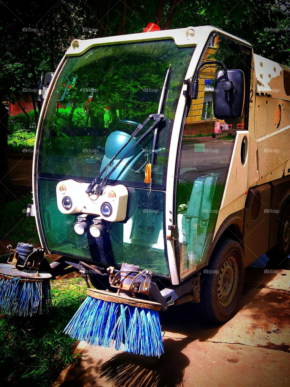 A subcompact road sweeper standing on a sidewalk against a backdrop of green trees