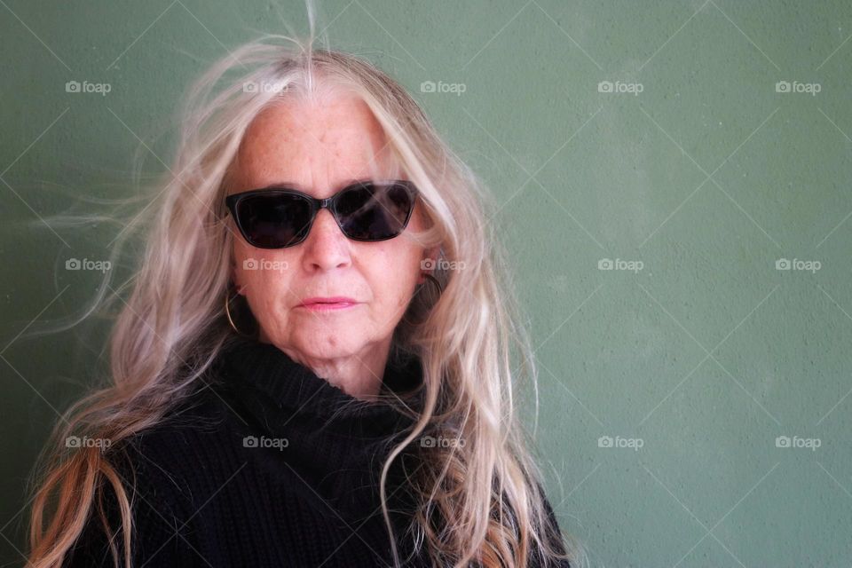 Portrait of a senior woman with long blonde blown hair, gold earrings and large sunglasses against a green background 