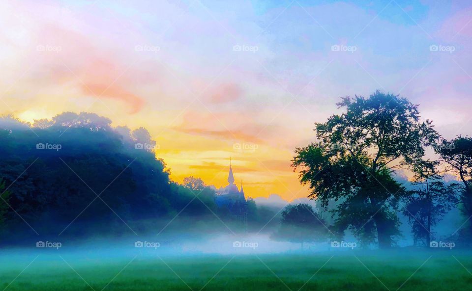 Castle between the trees in the distance, hidden behind a curtain of fog and with a background of a colorful sunrise 