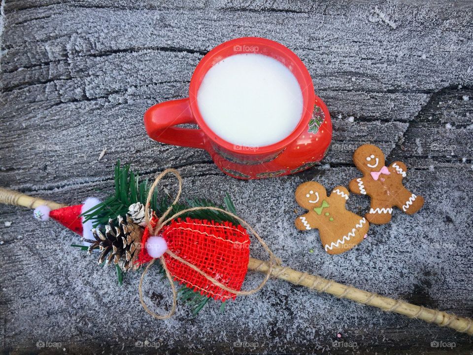 Red boot-shaped mug of white chocolate and milk on snowy table with two gingerbread men beside and frozen pine cone tree branches