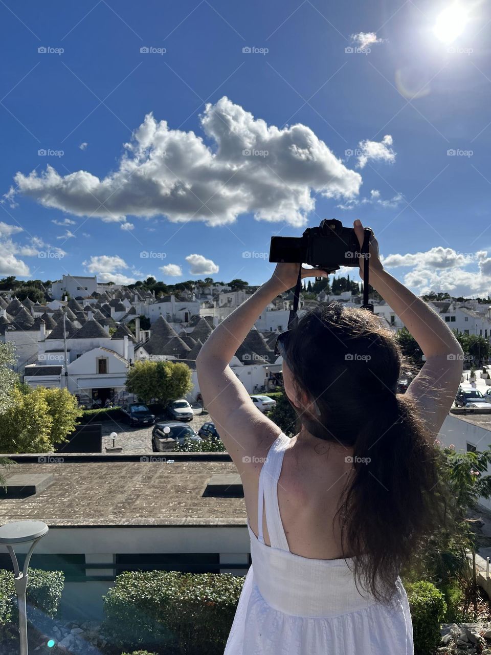 Portrait of one young Caucasian brunette girl standing from the back and photographing with a camera the ancient city of Alberobello in Italy on a sunny summer day, close-up side view.