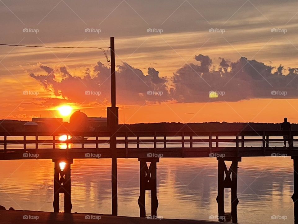 The sky is ablaze with a vivid shade of orange as the sun sets behind a rustic pier at Hampton Harbor in Hampton, NH. The silhouette of the pier stretches out into the water, adding a peaceful yet dramatic focal point to the scene.