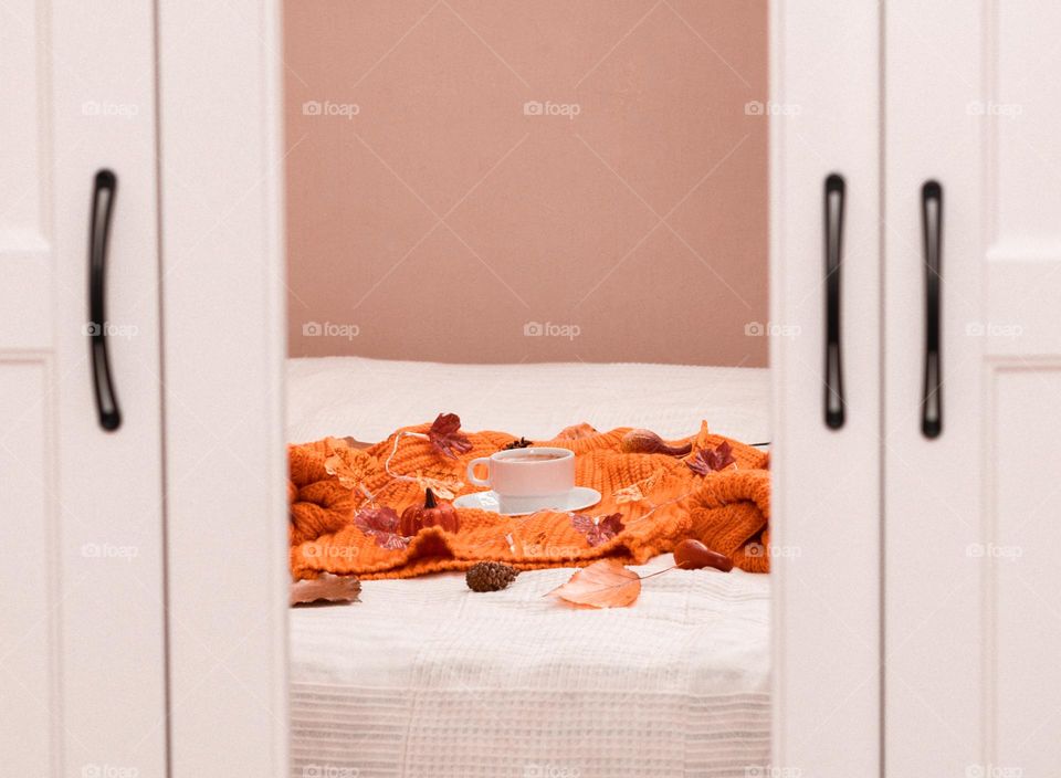 One cup of coffee with an orange knitted sweater, autumn leaves and a garland lie on a bed with a white bedspread and are reflected in the mirror of a white wardrobe, close-up side view.