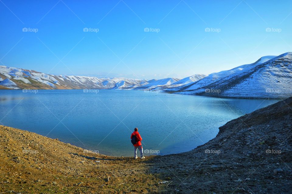 the traveler is standing against a huge mountain lake. clear spring sky. behind the lake you can see snow-covered hills and mountains.