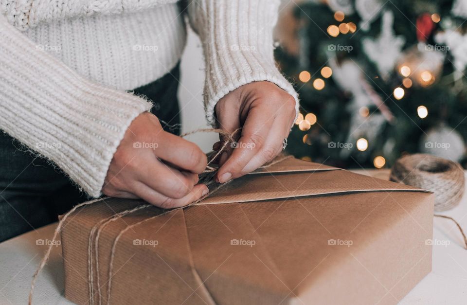 Midsection of woman wrapping christmas present in brown paper in front of christmas tree at home