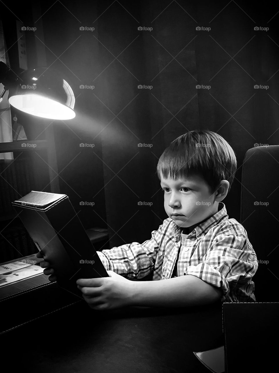 Close-up black-and-white portrait of a small child boy sitting at a table under a lamp holding a book in his hands