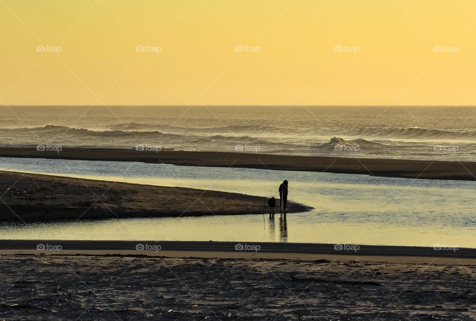 An adult and child explore pools of water on the beach just after sunset with the Atlantic Ocean rolling in the distance 