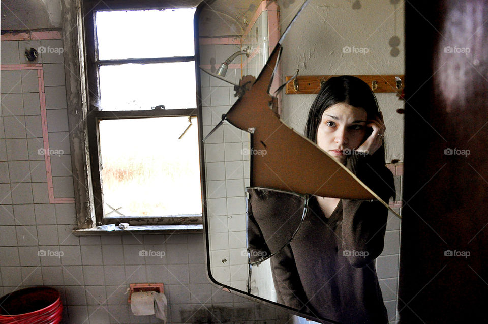 Young woman looks at her reflection in a broken mirror in an abandoned house.